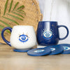 Two mugs with evil eye designs on a wooden surface with coasters.