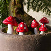 A set of whimsical, handcrafted mushroom plant markers with red caps and white stems, displayed in a pot with moss.