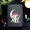 A5 notebook with a mystical mushroom design, gold foil page edges, and a ribbon marker, surrounded by small decorative items on a dark background.