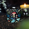 A black ceramic mug with a mystical forest print, featuring mushrooms, leaves, and flowers
