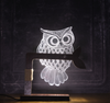 An illuminated owl table lamp with a branch-shaped wooden stand, featuring a lace-like texture on the owl's body, mounted on a dark surface.
