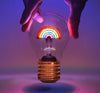 A hand reaching out to a cordless rainbow-colored lightbulb nightlight table lamp, illuminated with a neon glow.