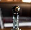 An 8-ball bottle stopper placed on a clear bottle, presumably sealing it. The background is blurred, featuring items that might be on a table such as books and a record.