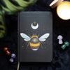 A5 notebook with a mystical bee design on the cover, accompanied by a ribbon marker, pens, and decorative stones on a dark textured background.