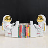 Two white astronaut bookends with gold visors and red detailing, playing a piano with books on it.