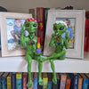 Two green alien figurines with peace signs in front of framed photos and books.