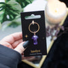 A hand holding a keyring with a mushroom-shaped amethyst gemstone attached to a gold keyring, presented on a black backing card with the text 'Amethyst Mushroom Keying'.