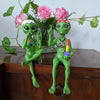 Two green alien figurines with accessories on a wooden surface with pink flowers in the background.