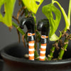 Two decorative plant pot ornaments designed to resemble spindly witch's legs with orange and white striped socks and black boots, placed in a plant pot with green foliage in the background.