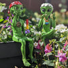 Two green alien figurines sitting outdoors among flowers with a colorful background