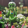 Green alien figurine with floral headband and peace sign gesture, surrounded by flowers.